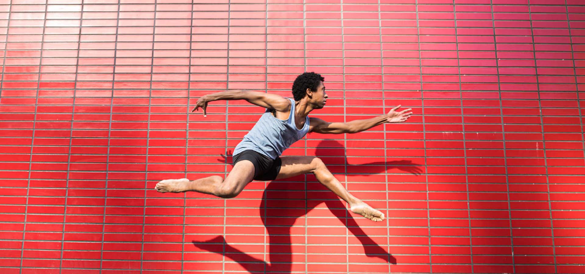 Male dancer doing a leap in front of a red tile building.